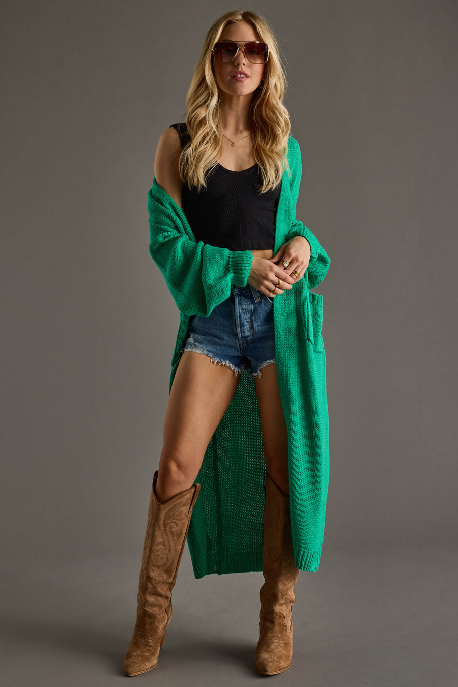 A woman wearing sunglasses, the Panache Apparel Collection Hilton Duster Cardigan in green over a black tank top, denim shorts, and tan knee-high boots stands against a neutral background.