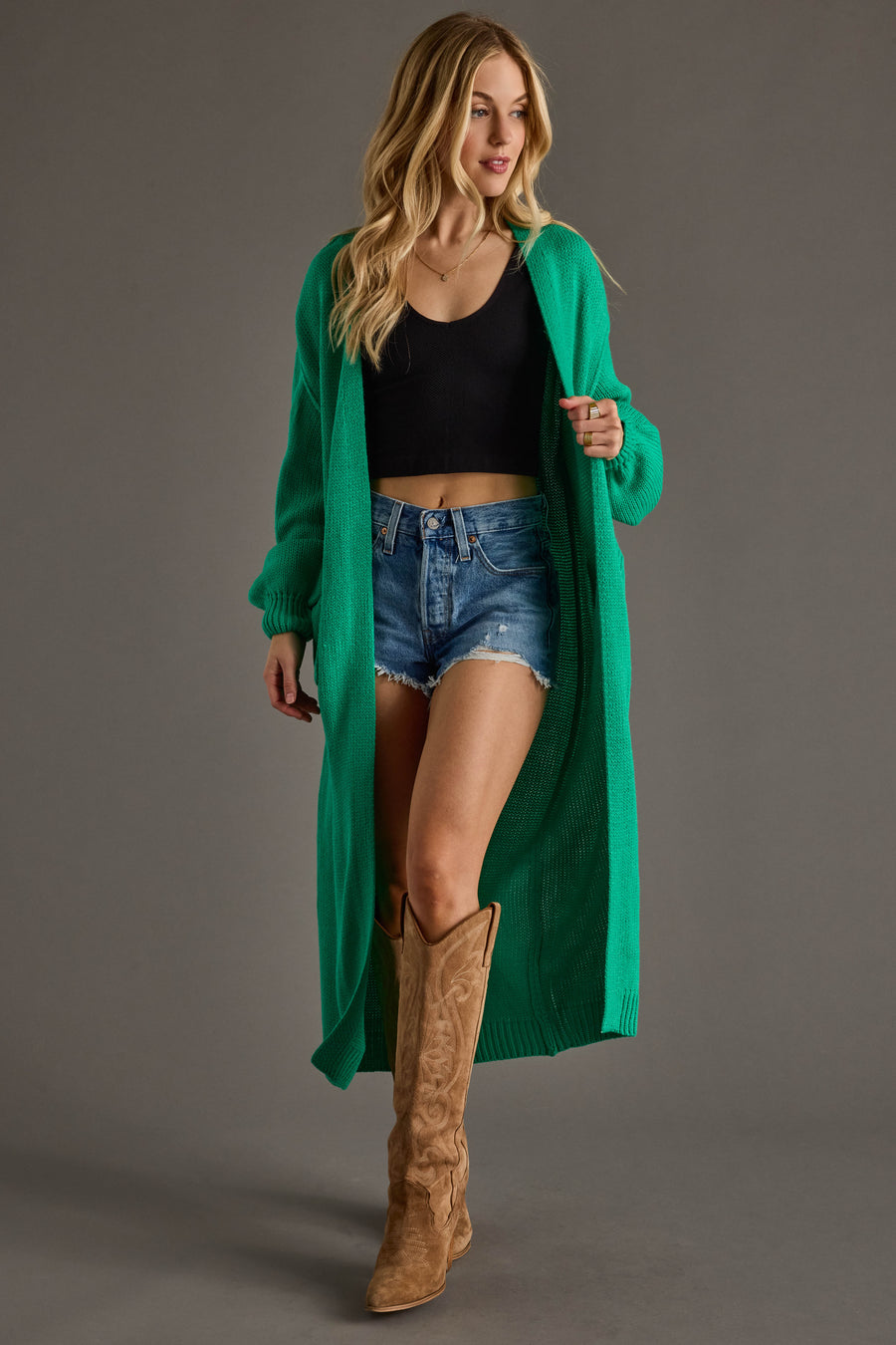 A woman models the Hilton Duster Cardigan in green from Panache Apparel Collection, paired with a black crop top, denim shorts, and brown cowboy boots against a gray background for a stylish layered look.
