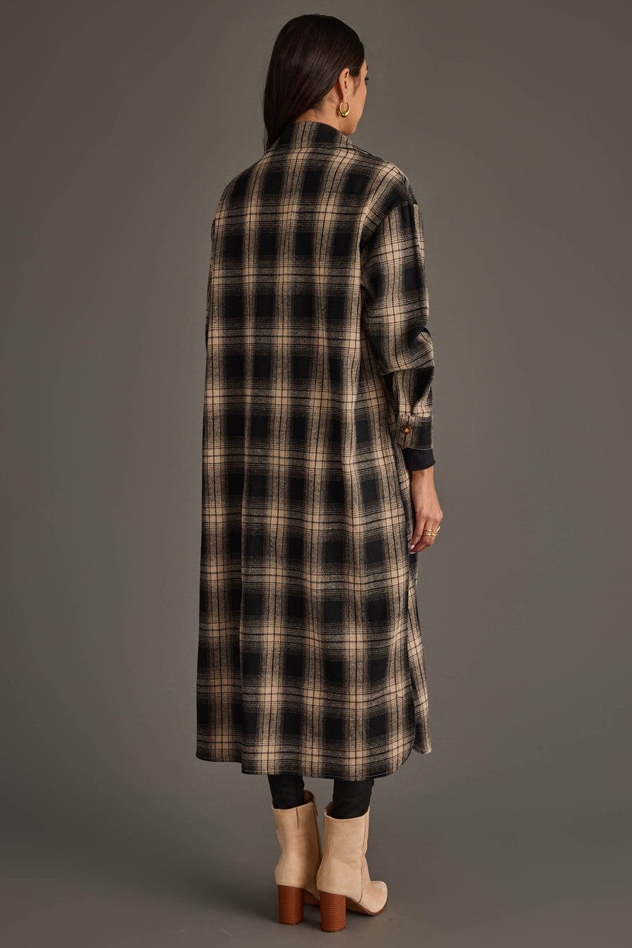 A woman stands with her back to the camera against a plain gray background, wearing the Panache Apparel Collection Black & Tan Plaid Long Lightweight Jacket, paired with black pants and tan heeled boots.