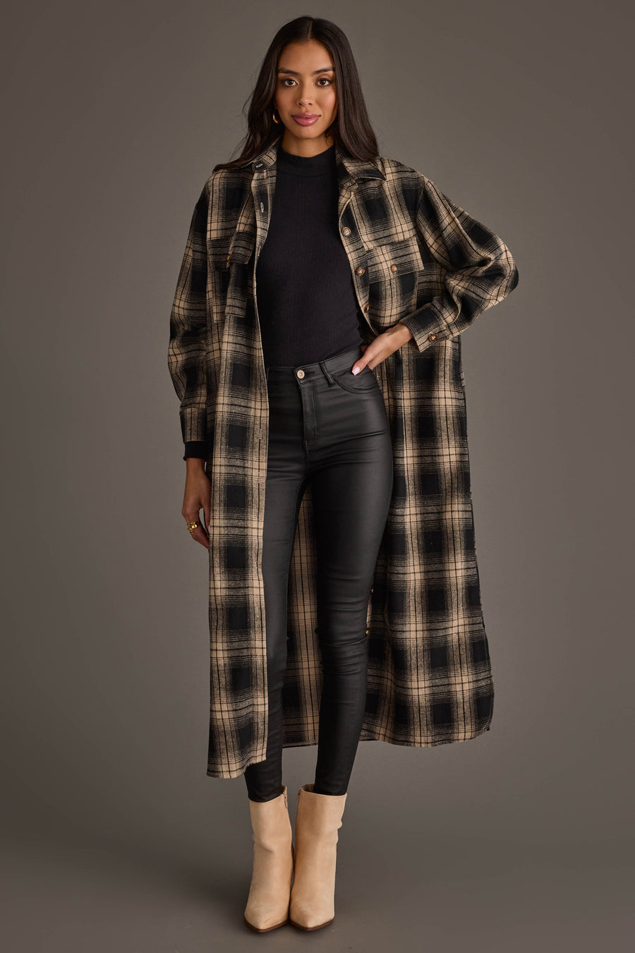 A woman is wearing the Panache Apparel Collection Black & Tan Plaid Long Lightweight Jacket over a black top and black skinny pants, paired with tan ankle boots, against a plain gray background.