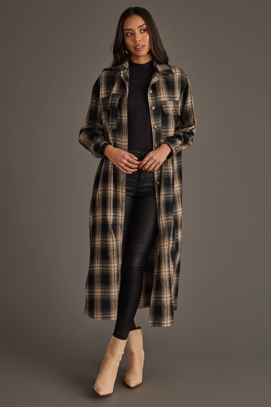 A woman with long dark hair and a neutral expression stands against a gray background, wearing the Panache Apparel Collection Black & Tan Plaid Long Lightweight Jacket over a black top and black pants, paired with beige ankle boots.