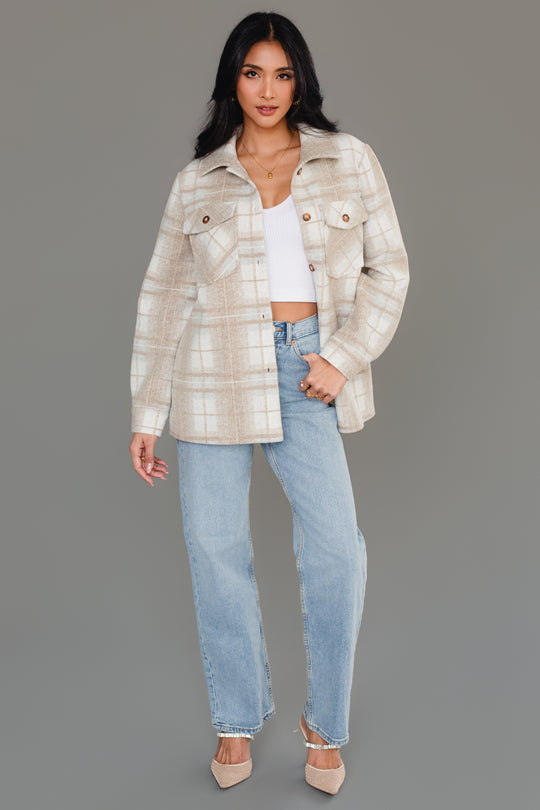 A woman with long dark hair stands in a relaxed, confident pose against a gray background, wearing the Panache Apparel Collection’s Tan, Cream & Gray Plaid Jacket over a white crop top, paired with light blue jeans and beige heels.