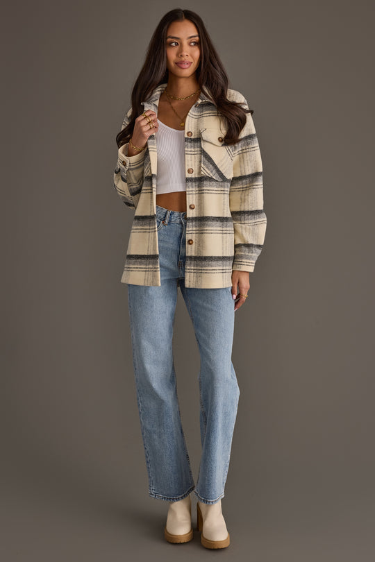 A woman stands against a gray background wearing the Panache Apparel Collection Cream, Black & Gray Plaid Jacket over a white crop top, paired with light blue jeans and beige heeled boots. She has long hair, layered necklaces, and smiles while touching her jacket.