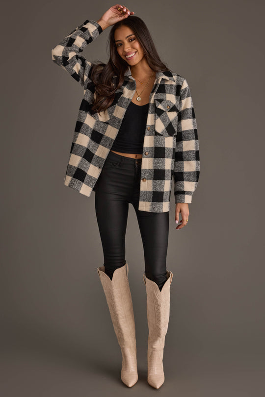 A woman wearing the Panache Apparel Collection Black & Cream Plaid Jacket over a black crop top, paired with black pants and beige cowboy boots, smiles and touches her hair while standing against a plain background.