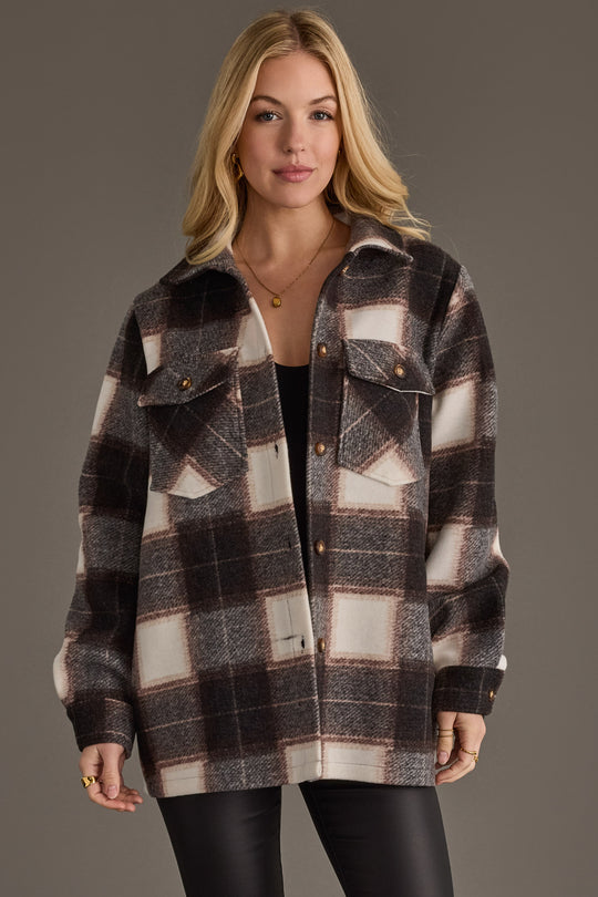 A woman with long blonde hair is wearing the Panache Apparel Collection Black, Brown & White Plaid Jacket over a black top and black pants, standing against a plain gray background.