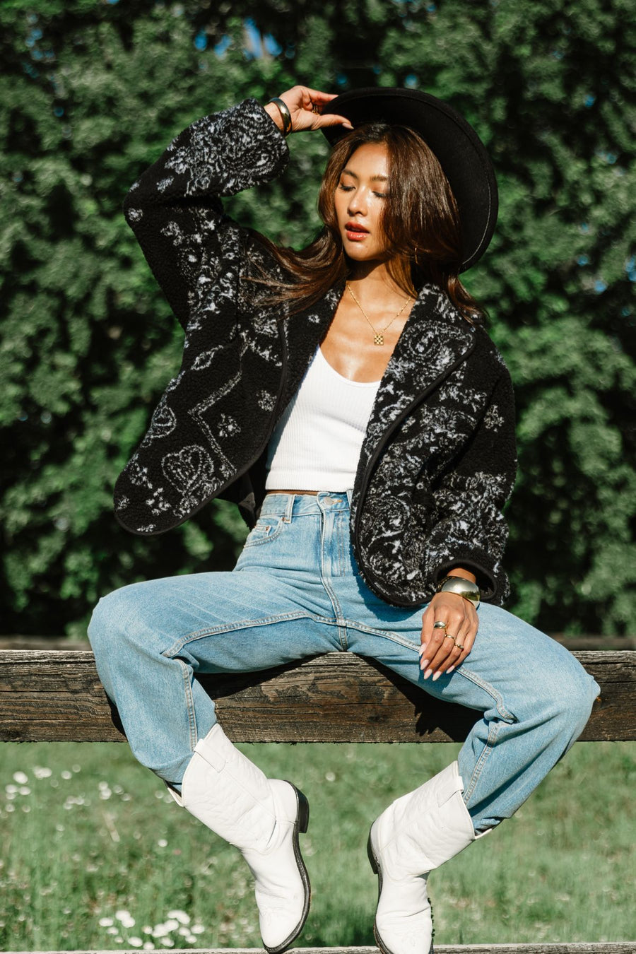 Outdoor lifestyle shot of a woman wearing Panache Apparel’s black and white paisley fleece coatigan with blue jeans and white western boots.