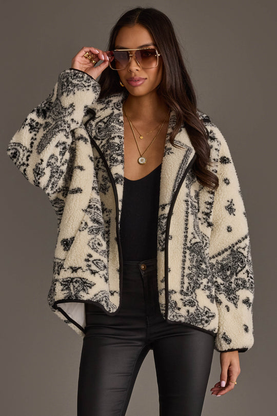 A woman wearing sunglasses, a black top, black leather pants, and the Panache Apparel Collection White & Black Paisley Fleece Coatigan poses against a plain gray background.