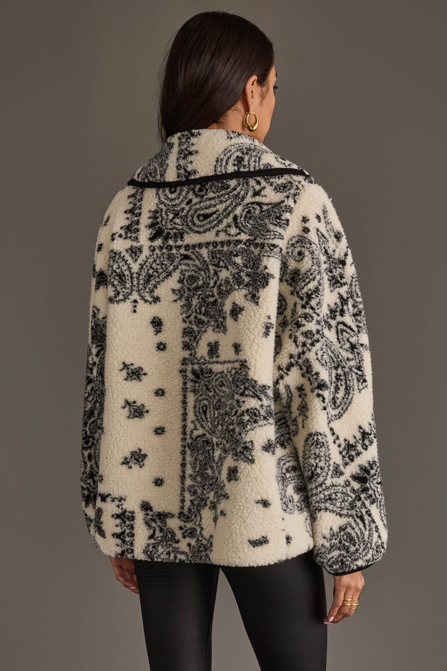 A woman with long dark hair models the Panache Apparel Collection White & Black Paisley Fleece Coatigan, facing away to highlight the intricate paisley pattern on the back. She accessorizes with gold hoop earrings and rings.