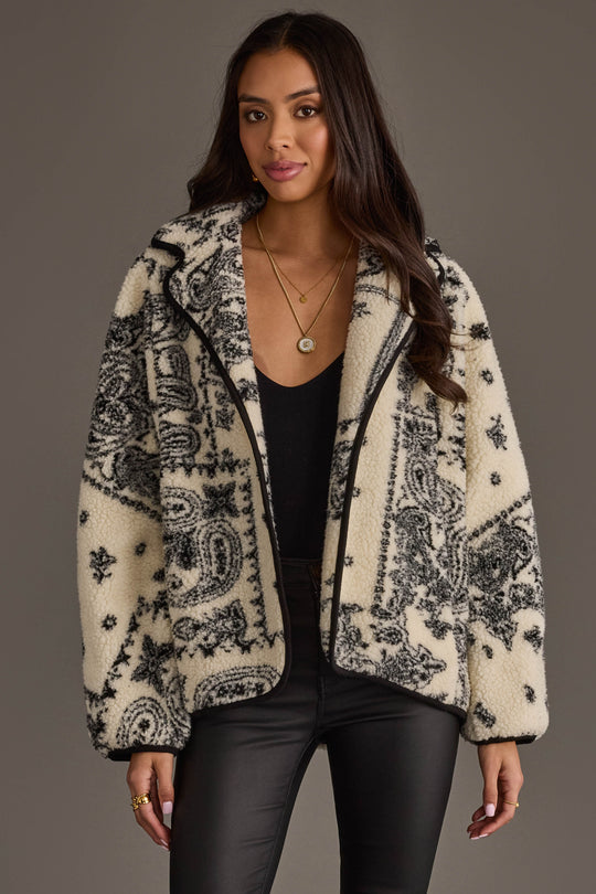 A woman wearing a black top, black leather pants, and the Panache Apparel Collection White & Black Paisley Fleece Coatigan stands against a plain background, looking at the camera with a slight smile.
