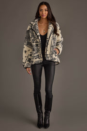 A woman stands against a neutral background wearing a black top, black leather pants, black boots, and the Panache Apparel Collection White & Black Paisley Fleece Coatigan with her hand in one pocket. She has long hair and accessorizes with necklaces and rings.