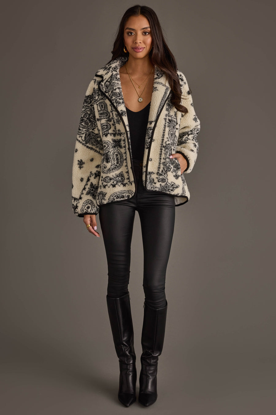 A woman stands against a neutral background wearing a black top, black leather pants, black boots, and the Panache Apparel Collection White & Black Paisley Fleece Coatigan with her hand in one pocket. She has long hair and accessorizes with necklaces and rings.