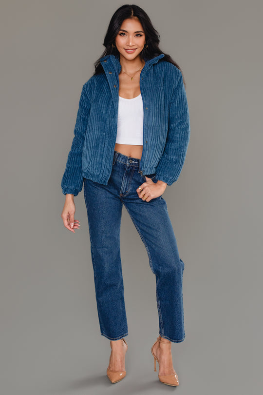 A woman poses against a plain gray background wearing the Panache Apparel Collection Blue Corduroy Puffer Jacket, paired with a white crop top, blue jeans, and nude heels.