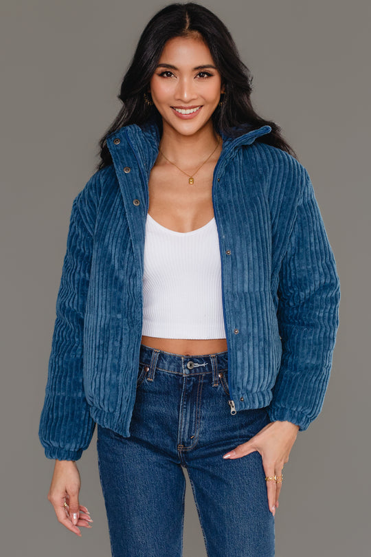 A woman smiles against a plain gray background wearing the Panache Apparel Collection Blue Corduroy Puffer Jacket layered over a white crop top and jeans.