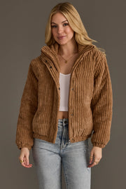 A woman wearing the Panache Apparel Collection’s Tan Corduroy Puffer Jacket over a white crop top and light blue jeans stands against a plain background, her expression relaxed and hands by her sides.