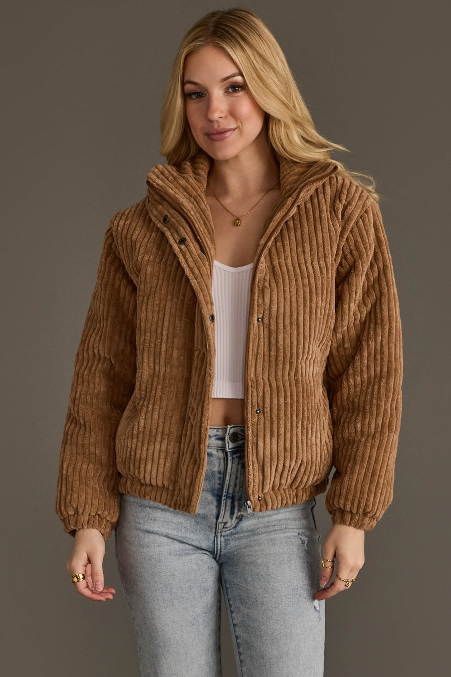 A woman wearing the Panache Apparel Collection’s Tan Corduroy Puffer Jacket over a white crop top and light blue jeans stands against a plain background, her expression relaxed and hands by her sides.