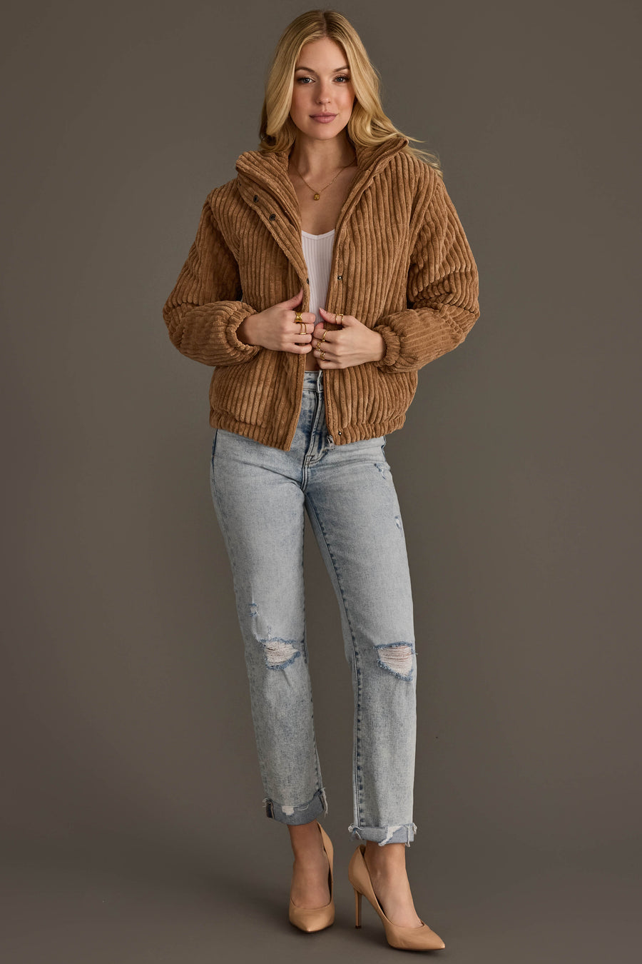 A person wearing the Tan Corduroy Puffer Jacket from Panache Apparel Collection, styled with a white top, light ripped jeans, and beige heels, stands against a plain gray background.