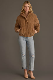 A woman smiles while wearing the Panache Apparel Collection Tan Corduroy Puffer Jacket, paired with light ripped jeans and nude heels, standing with her hands in the jacket pockets against a plain gray background.