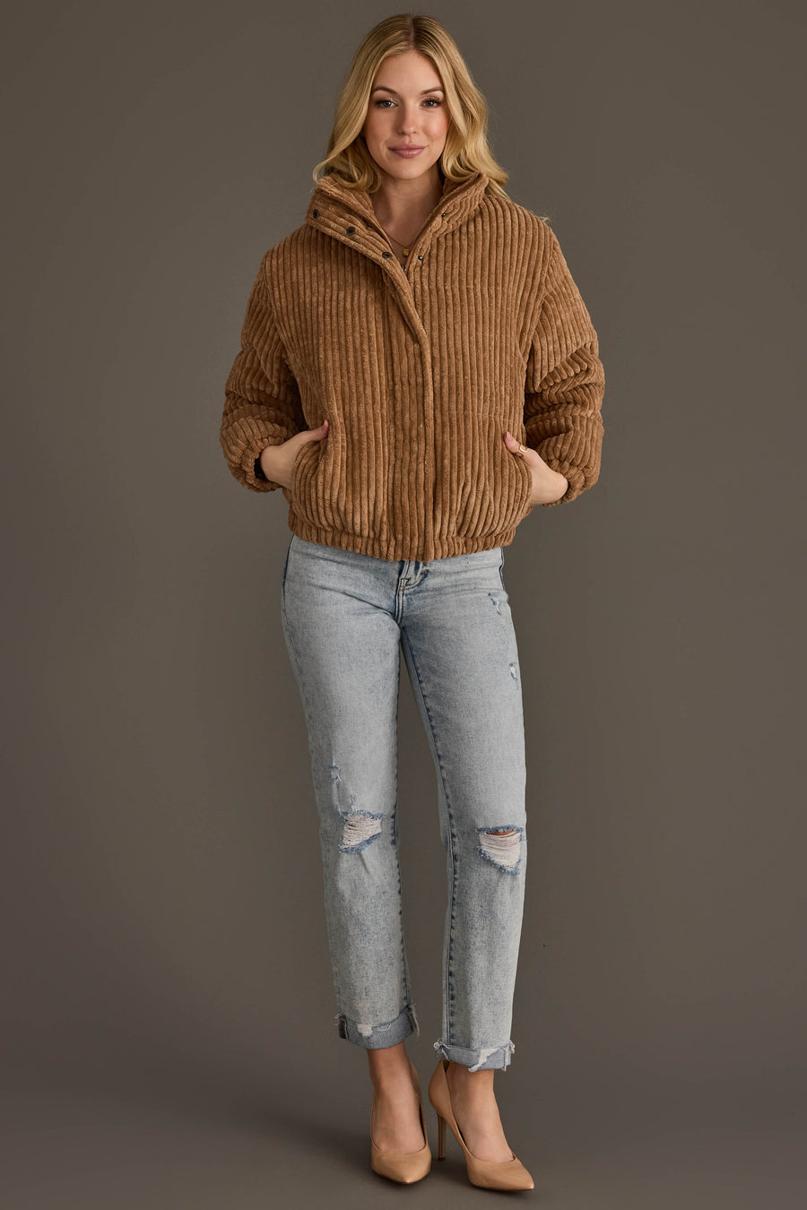 A woman smiles while wearing the Panache Apparel Collection Tan Corduroy Puffer Jacket, paired with light ripped jeans and nude heels, standing with her hands in the jacket pockets against a plain gray background.