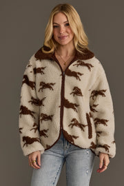 A woman models the Panache Apparel Collection Cream & Brown Fleece Horse Jacket, featuring a cozy cream-colored design with brown horse patterns, paired with light blue jeans and a gold necklace, as she smiles gently at the camera against a plain gray background.