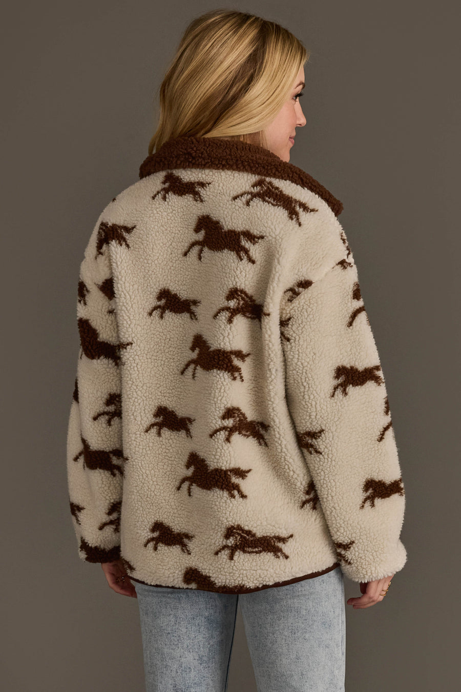A woman with blonde hair models the Panache Apparel Collection Cream & Brown Fleece Horse Jacket, styled with light blue jeans and posed against a plain gray background.