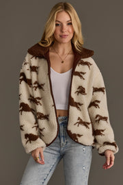 A woman wearing the Panache Apparel Collection Cream & Brown Fleece Horse Jacket over a white crop top and light blue jeans stands against a plain gray background, smiling softly.