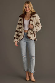 A woman with blonde hair styled in loose waves stands against a gray background, wearing a white crop top, the Cream & Brown Fleece Horse Jacket from Panache Apparel Collection, ripped light blue jeans, and beige heels.