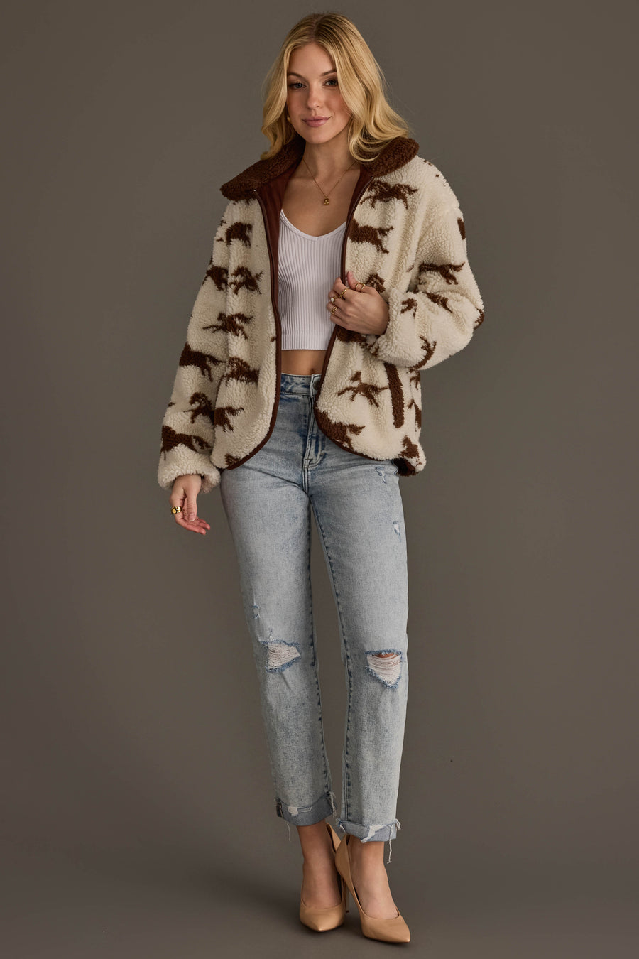 A woman with blonde hair styled in loose waves stands against a gray background, wearing a white crop top, the Cream & Brown Fleece Horse Jacket from Panache Apparel Collection, ripped light blue jeans, and beige heels.