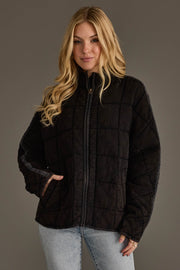 A woman with long blonde hair, dressed in the Panache Apparel Collection’s Faded Black Soft Quilted Jacket and light blue jeans, stands against a plain gray background with one hand in her pocket and a slight smile.