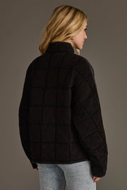 A woman with long blonde hair is seen from behind, standing against a plain gray background and wearing the Faded Black Soft Quilted Jacket by Panache Apparel Collection along with light blue jeans.