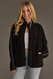 A woman with long blonde hair, dressed in the Panache Apparel Collection’s Faded Black Soft Quilted Jacket over a white top and light blue jeans, stands smiling against a plain gray background.