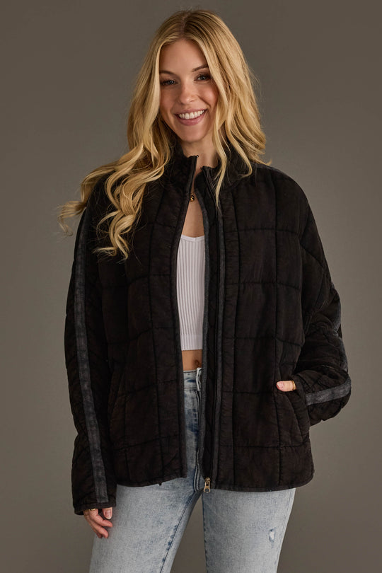 A woman with long blonde hair, dressed in the Panache Apparel Collection’s Faded Black Soft Quilted Jacket over a white top and light blue jeans, stands smiling against a plain gray background.