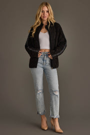 A woman stands against a gray background, wearing the Panache Apparel Collection’s Faded Black Soft Quilted Jacket, paired with a white ribbed crop top, light blue ripped jeans, and beige high heels. She has long blonde hair and is smiling slightly.