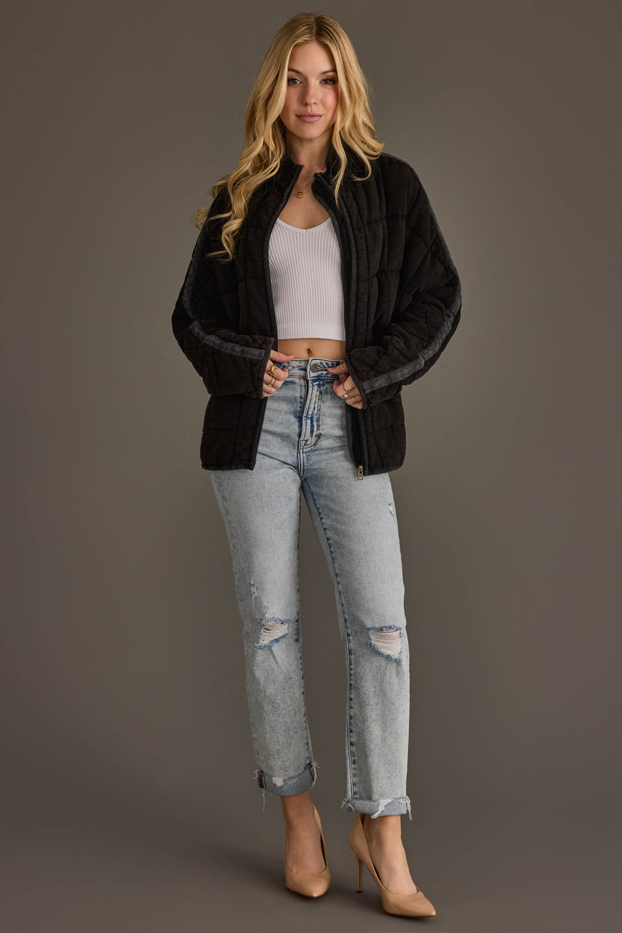 A woman stands against a gray background, wearing the Panache Apparel Collection’s Faded Black Soft Quilted Jacket, paired with a white ribbed crop top, light blue ripped jeans, and beige high heels. She has long blonde hair and is smiling slightly.