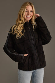 A woman with long blonde hair wears the Panache Apparel Collection’s Faded Black Soft Quilted Jacket and light blue jeans, smiling and posing with one hand touching her hair against a plain background.