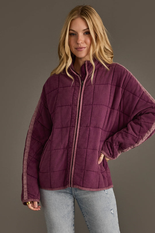 A woman wearing the Panache Apparel Collection’s Plum Soft Quilted Jacket and light blue jeans stands against a plain gray background, smiling with one hand on her hip.