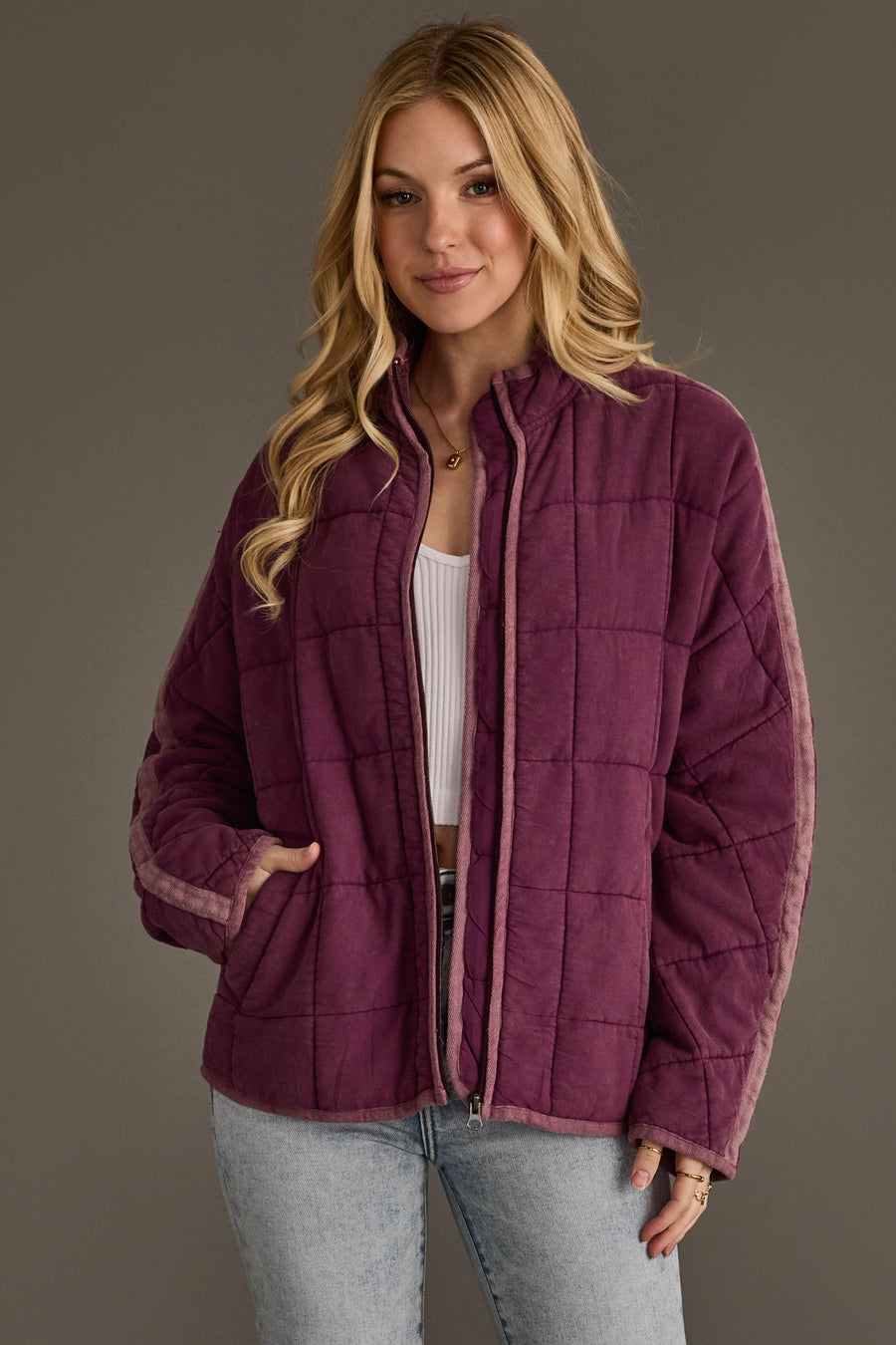 A woman with long blonde hair wears the Plum Soft Quilted Jacket from Panache Apparel Collection over a white top and light blue jeans, standing against a plain gray background.