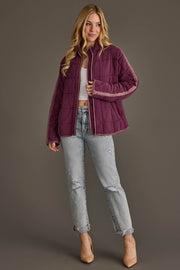 A woman with long blonde hair smiles slightly while posing against a gray background, dressed in the Panache Apparel Collection Plum Soft Quilted Jacket layered over a white crop top, paired with light ripped jeans and beige heels.