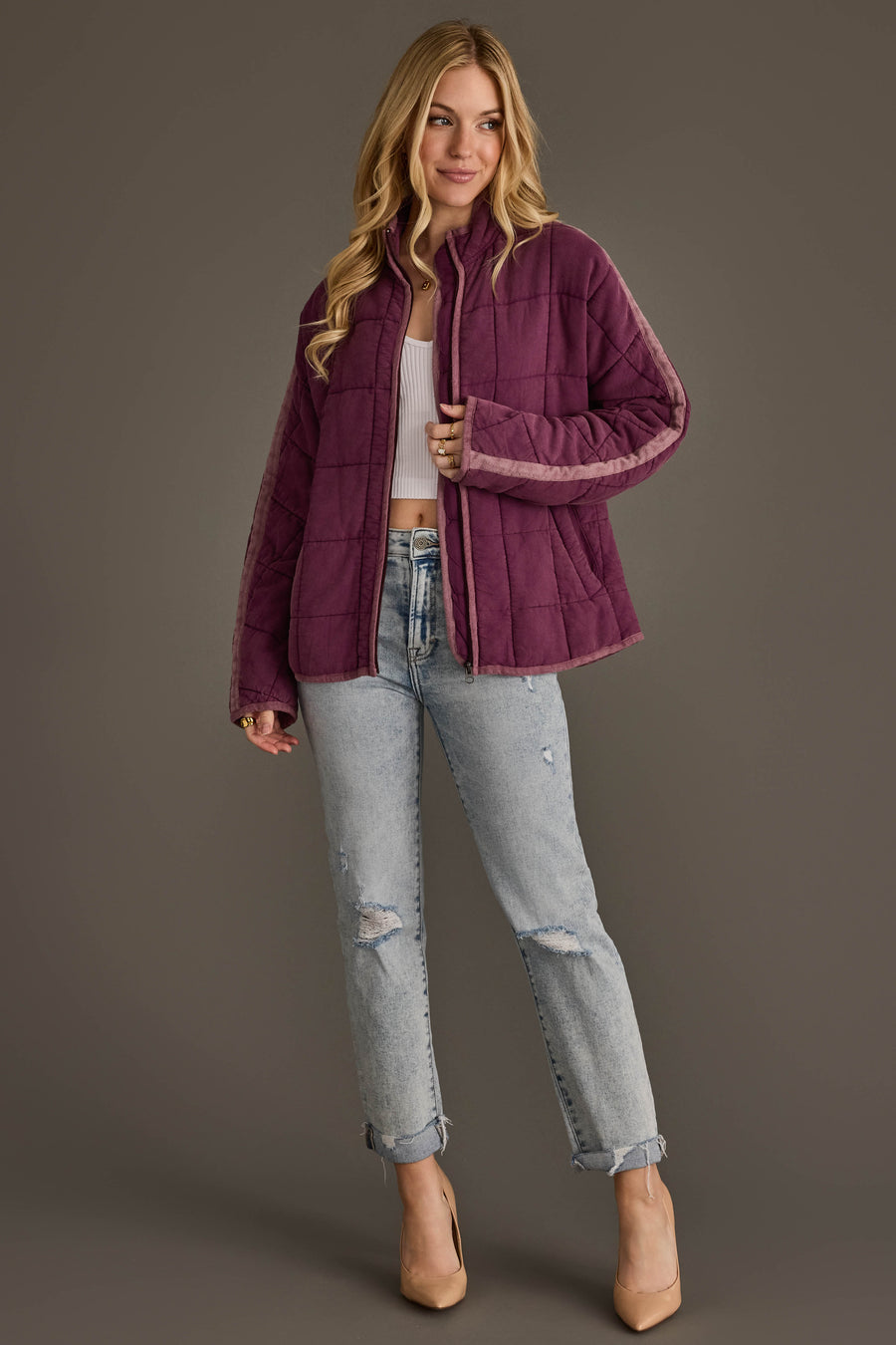 A woman with long blonde hair smiles slightly while posing against a gray background, dressed in the Panache Apparel Collection Plum Soft Quilted Jacket layered over a white crop top, paired with light ripped jeans and beige heels.