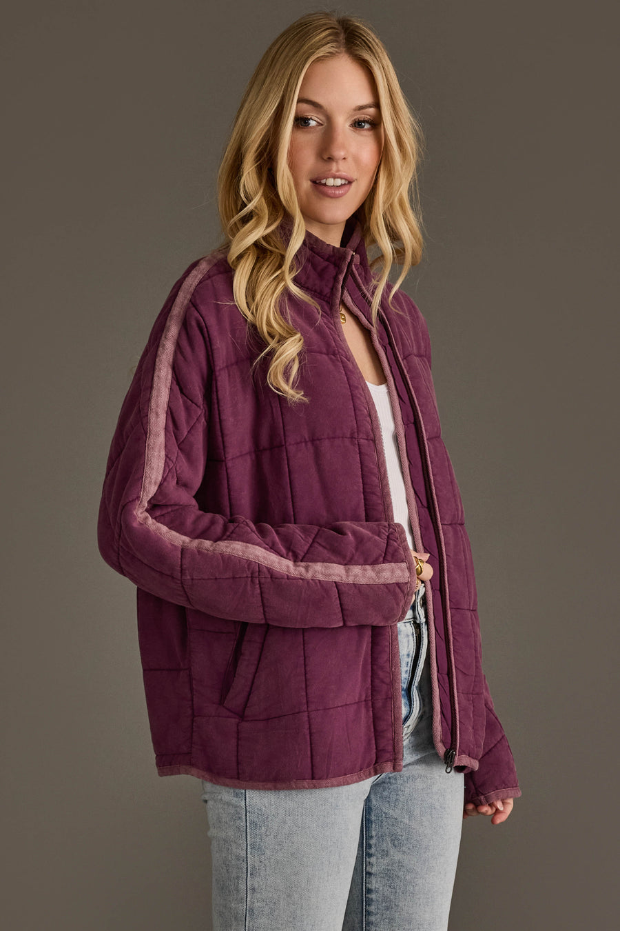 A woman wearing the Plum Soft Quilted Jacket from the Panache Apparel Collection, featuring light pink stripes on the sleeves, paired with a white top and light blue jeans, stands against a plain gray background.