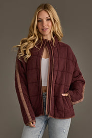 A woman with long blonde hair is seen wearing the Panache Apparel Collection’s Dark Brown Soft Quilted Jacket, paired with a white ribbed crop top and light blue jeans, standing against a plain gray background.