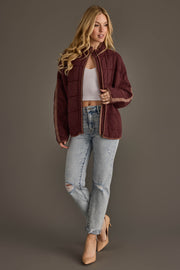 A woman with long blonde hair wears the Panache Apparel Collection’s Dark Brown Soft Quilted Jacket, a white crop top, ripped light blue jeans, and nude heels, standing against a plain gray background.