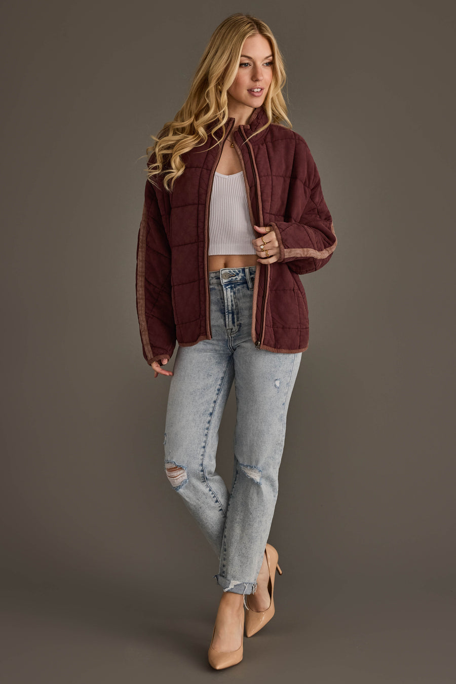 A woman with long blonde hair wears the Panache Apparel Collection’s Dark Brown Soft Quilted Jacket, a white crop top, ripped light blue jeans, and nude heels, standing against a plain gray background.