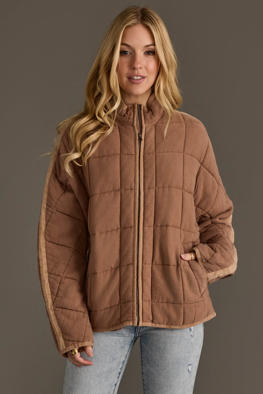 A woman with long blonde hair stands against a plain gray background, wearing the Panache Apparel Collection Tan Soft Quilted Jacket and light blue jeans, with one hand in her pocket.