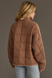 A woman with blonde hair stands with her back to the camera against a plain background, wearing the Tan Soft Quilted Jacket from Panache Apparel Collection and light blue jeans.