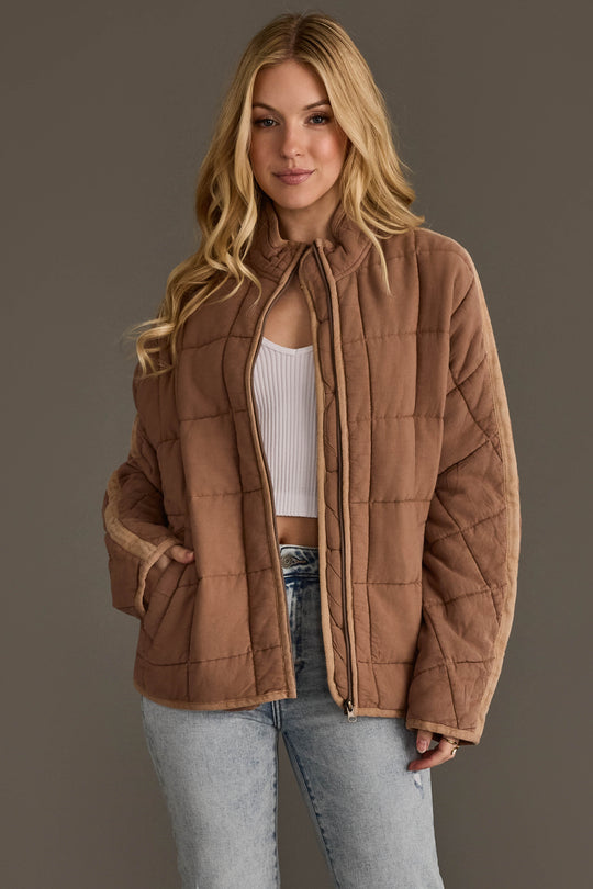 A woman wearing the Panache Apparel Collection Tan Soft Quilted Jacket over a white crop top and light blue jeans, standing against a plain gray background.