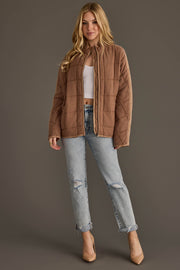 A woman wearing the Panache Apparel Collection Tan Soft Quilted Jacket stands against a plain brown background, styled with a white ribbed crop top, light blue ripped jeans, and beige heels.