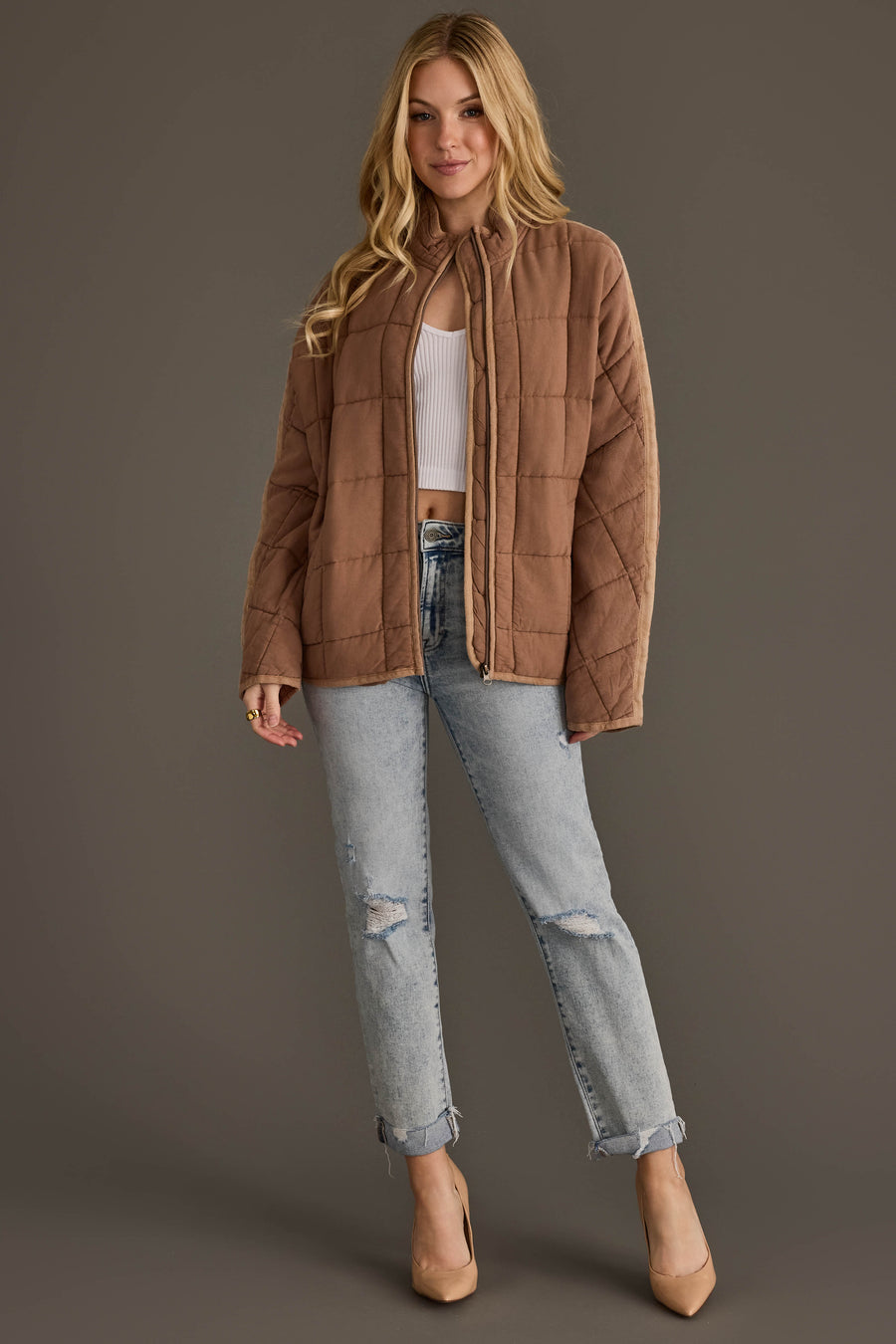 A woman wearing the Panache Apparel Collection Tan Soft Quilted Jacket stands against a plain brown background, styled with a white ribbed crop top, light blue ripped jeans, and beige heels.