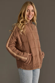 A smiling woman with long blonde hair showcases the Panache Apparel Collection’s Tan Soft Quilted Jacket, featuring beige stripes on the sleeves, paired with light blue jeans and set against a plain gray background.