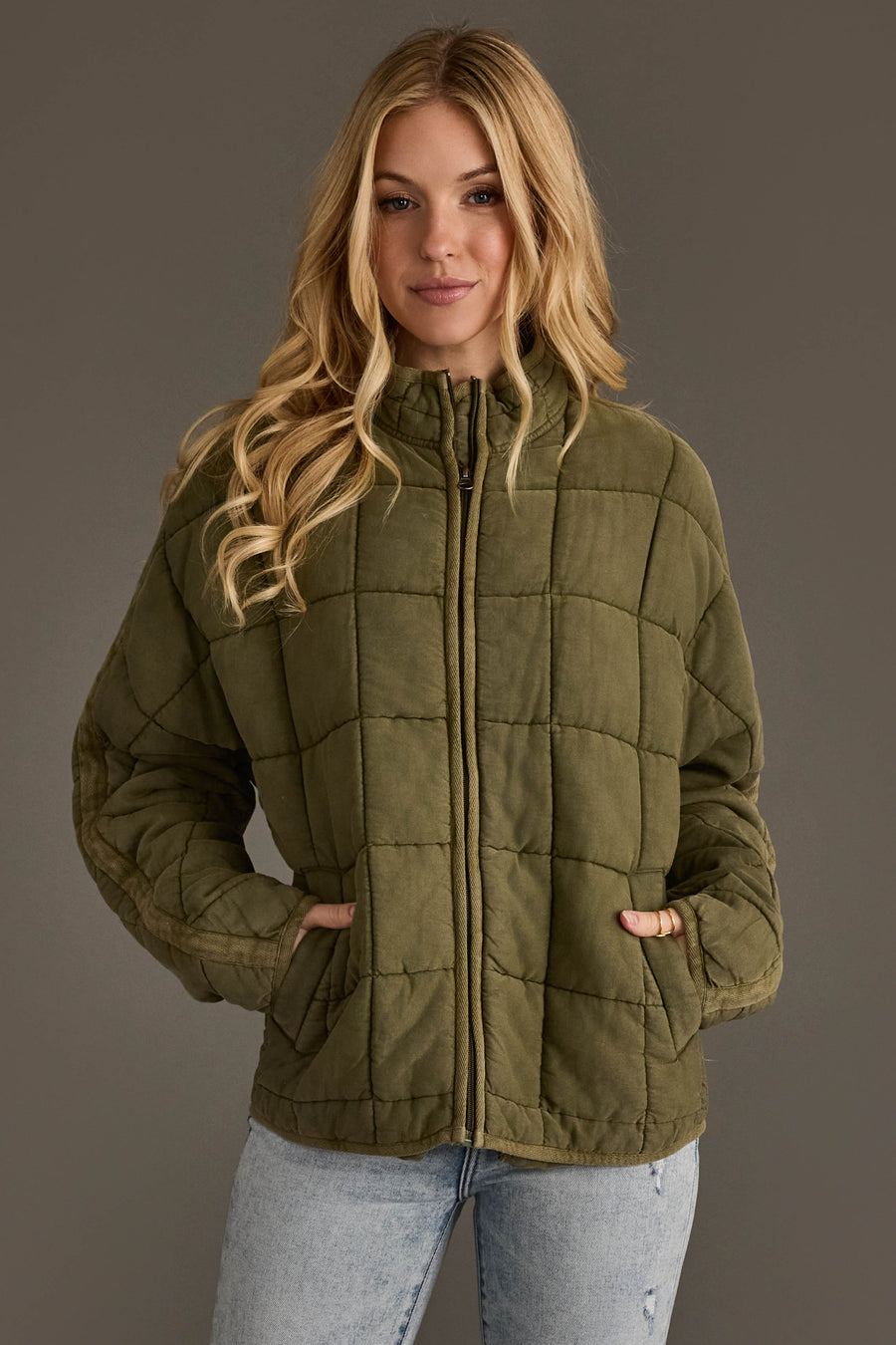 A person with long blonde hair is wearing the Olive Soft Quilted Jacket from Panache Apparel Collection and light blue jeans, standing against a plain gray background and looking at the camera with their hands in the jacket pockets.