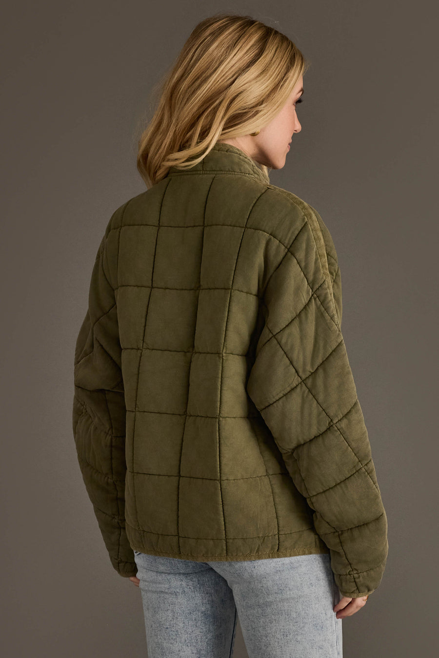 A person with blonde hair wears the Panache Apparel Collection Olive Soft Quilted Jacket and light blue jeans, standing against a plain gray background and facing away from the camera.